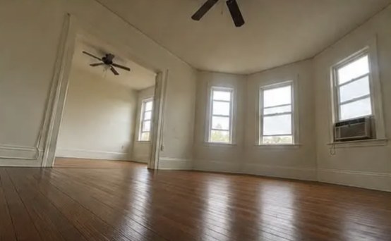 Empty room with hardwood floors, three tall windows in a bay, a ceiling fan, and a wide doorway into a second bright area